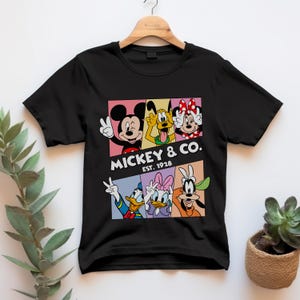 Puede incluir: Camiseta negra con un gr&aacute;fico colorido de Mickey Mouse, Pluto, Minnie Mouse, Donald Duck, Daisy Duck y Goofy. El gr&aacute;fico incluye el texto "Mickey & Co. Est. 1928". La camiseta est&aacute; colgada en una percha de madera.