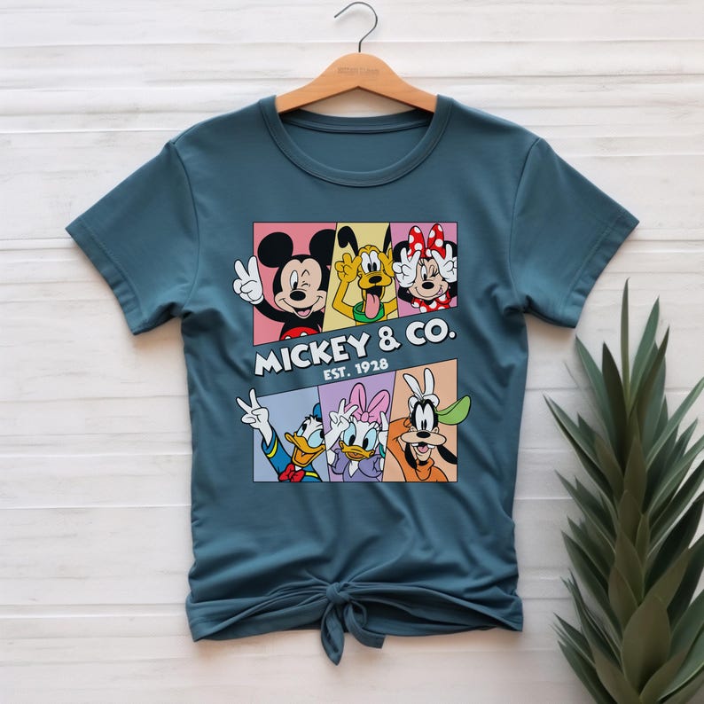 Puede incluir: Camiseta de color turquesa con un dise&ntilde;o colorido de Mickey Mouse, Pluto, Minnie Mouse, Donald Duck, Daisy Duck y Goofy. El dise&ntilde;o incluye el texto "Mickey & Co. Est. 1928". La camiseta est&aacute; colgada en una percha de madera.