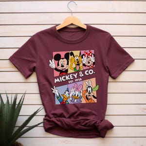 Puede incluir: Camiseta burdeos con un dise&ntilde;o colorido de Mickey Mouse, Pluto, Minnie Mouse, Donald Duck, Daisy Duck y Goofy. El texto "Mickey & Co. Est. 1928" est&aacute; impreso debajo.