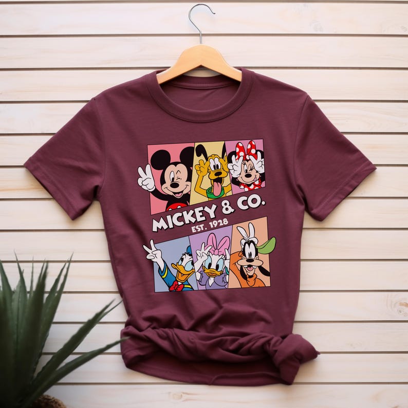Puede incluir: Camiseta burdeos con un dise&ntilde;o colorido de Mickey Mouse, Pluto, Minnie Mouse, Donald Duck, Daisy Duck y Goofy. El texto "Mickey & Co. Est. 1928" est&aacute; impreso debajo.