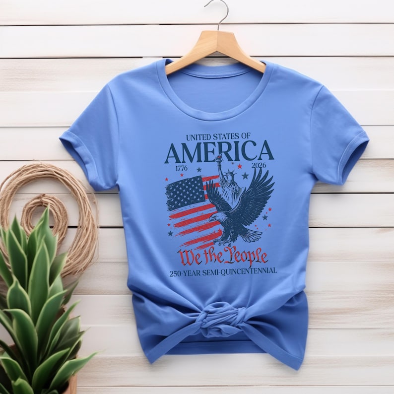 May include: A light blue t-shirt with a patriotic design. The graphic includes the American flag, an eagle, and the text "UNITED STATES OF AMERICA 1776 2026 We the People 250 YEAR SEMI-QUINCENTENNIAL". The shirt is knotted at the waist.