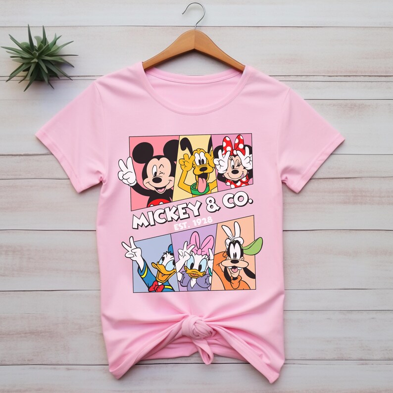 Puede incluir: Camiseta rosa claro con un dise&ntilde;o de Mickey Mouse, Pluto, Minnie Mouse, Donald Duck, Daisy Duck y Goofy. El dise&ntilde;o est&aacute; en un dise&ntilde;o rectangular con el texto "Mickey & Co. Est. 1928". La camiseta est&aacute; colgada en una percha de madera.