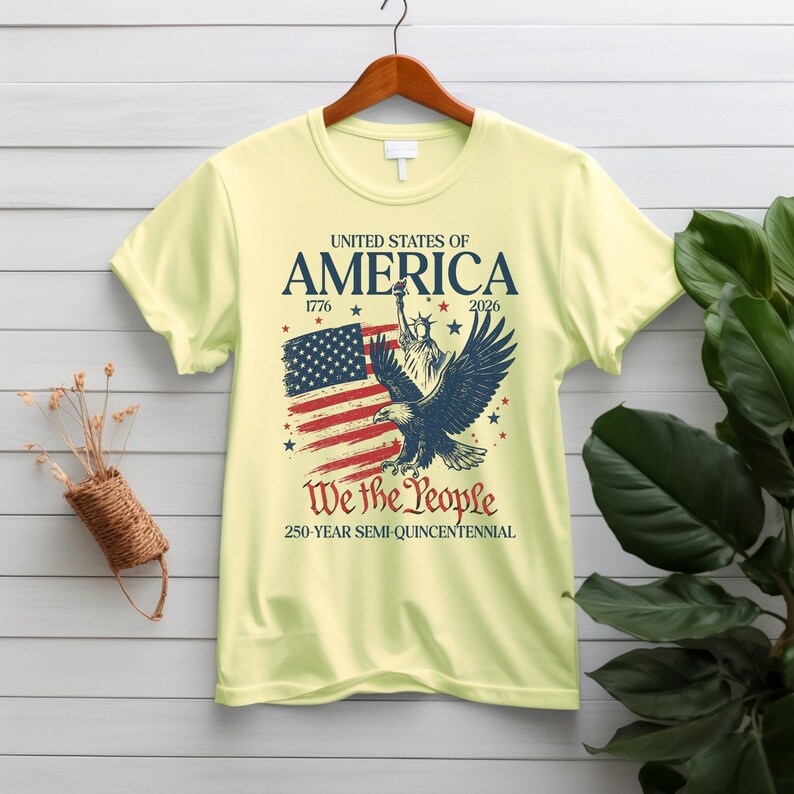 May include: A pale yellow t-shirt featuring a patriotic design. The design includes the text "UNITED STATES OF AMERICA 1776-2026" above an illustration of the American flag, the Statue of Liberty, and an eagle. The text "We the People" is below the illustration.