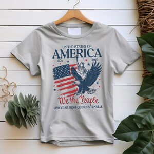 May include: A light grey t-shirt with a patriotic design. The design features the text "UNITED STATES OF AMERICA" above an American flag, the Statue of Liberty, and an eagle. The text "We the People" and "250-YEAR SEMI-QUINCENTENNIAL" are also included.