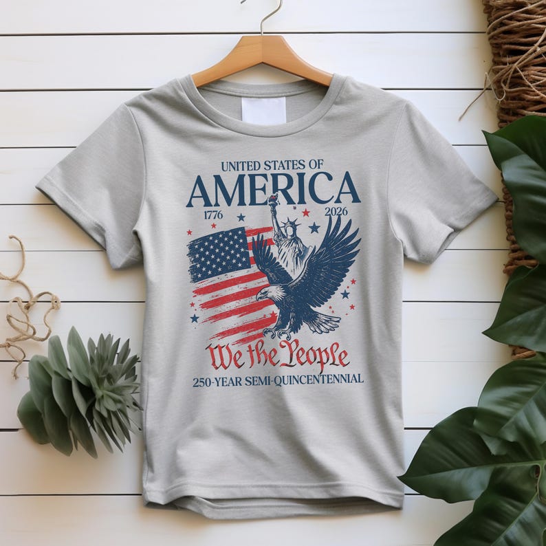 May include: A light grey t-shirt with a patriotic design. The design features the text "UNITED STATES OF AMERICA" above an American flag, the Statue of Liberty, and an eagle. The text "We the People" and "250-YEAR SEMI-QUINCENTENNIAL" are also included.