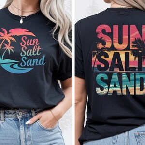 K&ouml;nnte beinhalten: Schwarzes T-Shirt mit einer farbenfrohen Grafik mit Palmen, einem Sonnenuntergang und den Worten "Sun Salt Sand". Das Design wiederholt sich auf der R&uuml;ckseite. Das Shirt besteht aus einem weichen Material.
