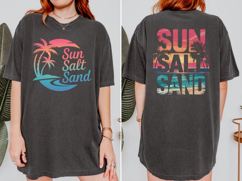 K&ouml;nnte beinhalten: Dunkelgraues T-Shirt mit einem farbenfrohen Grafikdesign eines Sonnenuntergangs, Palmen und den Worten "Sun Salt Sand". Das Design wiederholt sich auf der R&uuml;ckseite. Das Shirt hat kurze &Auml;rmel und eine lockere Passform.
