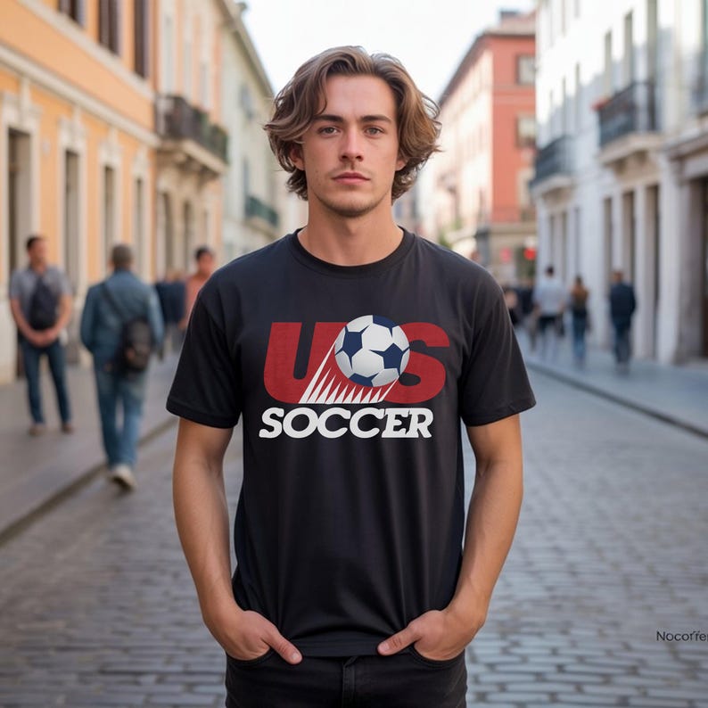 May include: A black short-sleeved t-shirt featuring a graphic print of a soccer ball and the red text US above the white text SOCCER. The design includes a motion effect behind the ball, centered on the chest of the garment.