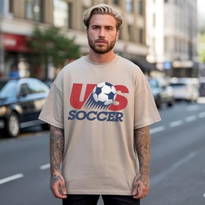 May include: A beige short-sleeved t-shirt featuring a graphic print on the chest. The design includes the large red letters US with a black and white soccer ball positioned over the S, and the word SOCCER printed in bold blue capital letters below.