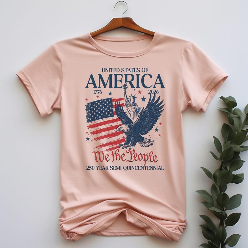 May include: A pale pink t-shirt with a patriotic design. The graphic features the American flag, the Statue of Liberty, and an eagle, with the text "UNITED STATES OF AMERICA 1776-2026" and "We the People 250-Year Semi-Quincentennial."