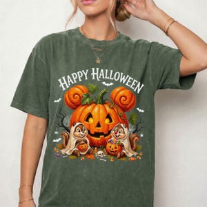 May include: A dark olive green t-shirt featuring a graphic print with the text Happy Halloween. The design includes a large orange jack-o'-lantern with mouse-ear-shaped pumpkins, two chipmunks wearing ghost costumes, and small bats.