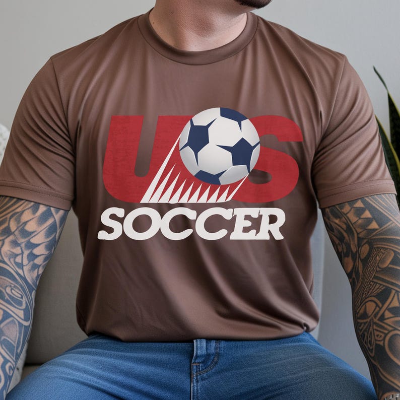 May include: A brown short-sleeved t-shirt featuring a graphic design with the red text US, a white and blue soccer ball positioned over the letters, and the word SOCCER printed in large white capital letters underneath the graphic.