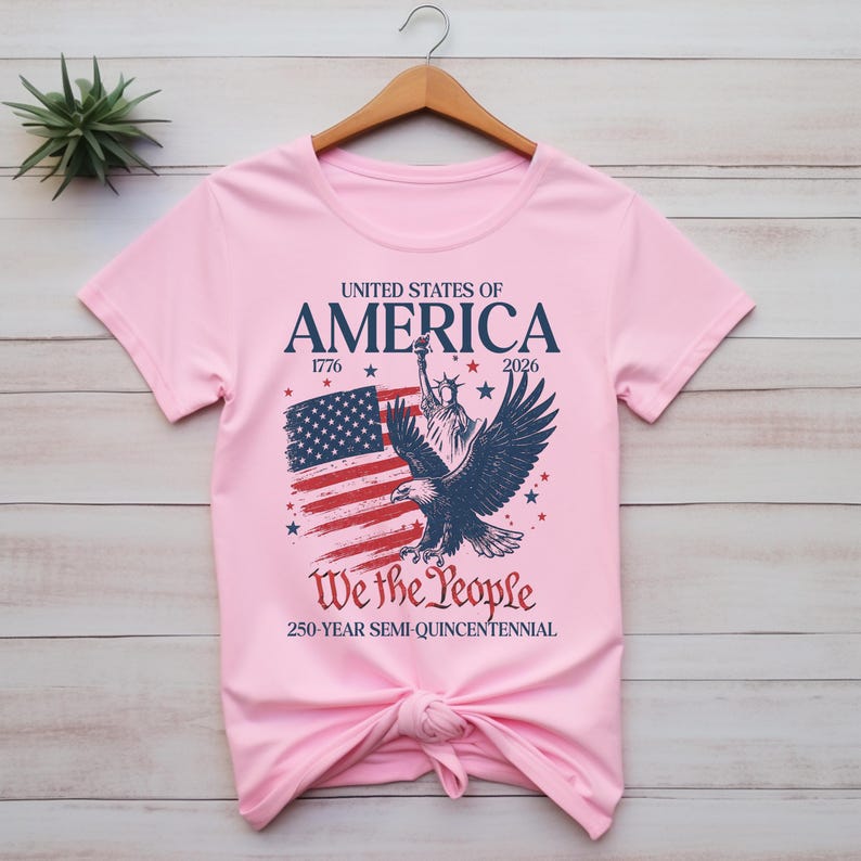 May include: A pale pink t-shirt with a patriotic design. The graphic includes the text "United States of America" with the years 1776 and 2026, an American flag, the Statue of Liberty, and an eagle. The text "We the People" is also included.