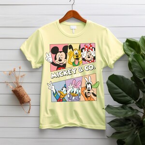 Puede incluir: Una camiseta amarilla clara con un gr&aacute;fico colorido de Mickey Mouse, Pluto, Minnie Mouse, Pato Donald, Daisy y Goofy. El texto "MICKEY & CO. EST. 1928" se muestra debajo. La camiseta est&aacute; colgada en una percha de madera.