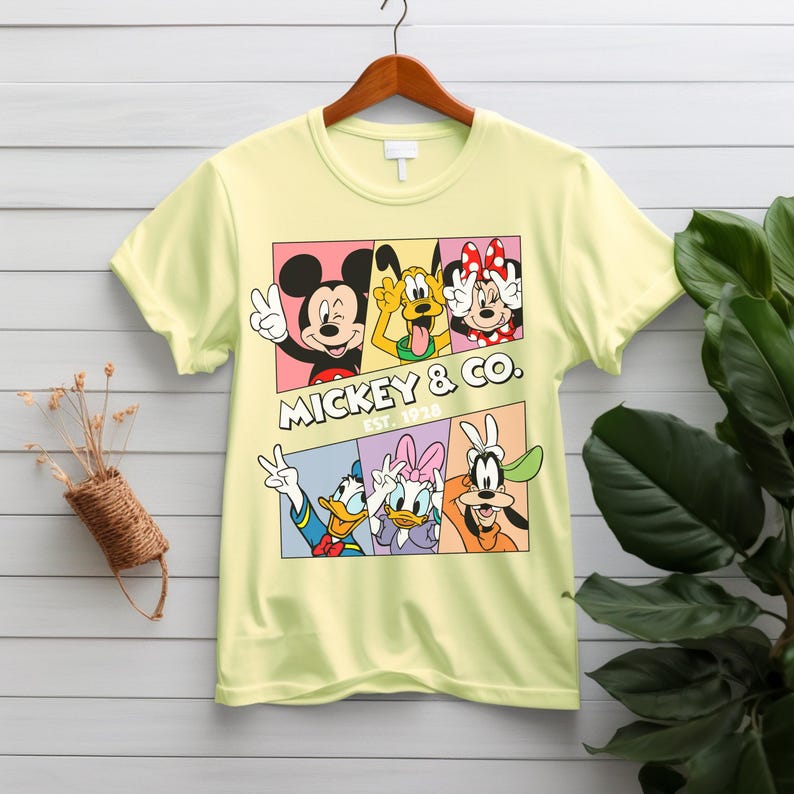 Puede incluir: Una camiseta amarilla clara con un gr&aacute;fico colorido de Mickey Mouse, Pluto, Minnie Mouse, Pato Donald, Daisy y Goofy. El texto "MICKEY & CO. EST. 1928" se muestra debajo. La camiseta est&aacute; colgada en una percha de madera.