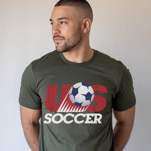 May include: An olive green short-sleeved crew neck t-shirt featuring a graphic design on the chest. The design includes the red letters US with a soccer ball positioned over the S, and the word SOCCER printed in white capital letters below.