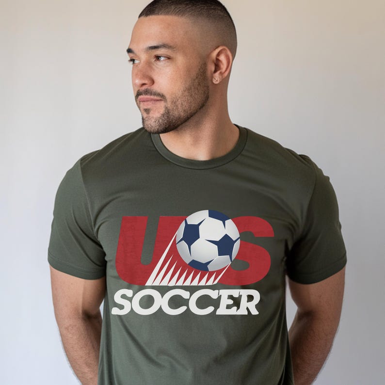 May include: An olive green short-sleeved crew neck t-shirt featuring a graphic design on the chest. The design includes the red letters US with a soccer ball positioned over the S, and the word SOCCER printed in white capital letters below.