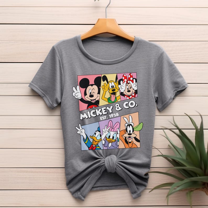 Puede incluir: Camiseta gris con un gr&aacute;fico colorido de Mickey Mouse, Pluto, Minnie Mouse, Donald Duck, Daisy Duck y Goofy. El gr&aacute;fico incluye el texto "Mickey & Co. Est. 1928". La camiseta est&aacute; colgada en una percha de madera.