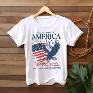 May include: White t-shirt with a patriotic design. The design features the text "UNITED STATES OF AMERICA", the American flag, the Statue of Liberty, and an eagle. The text "We the People" and "250-YEAR SEMI-QUINCENTENNIAL" are also included.