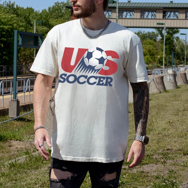 May include: A cream-colored short-sleeve t-shirt featuring a graphic on the chest with the red letters US, a black and white soccer ball replacing the letter O, and the word SOCCER printed in blue capital letters underneath the graphic.