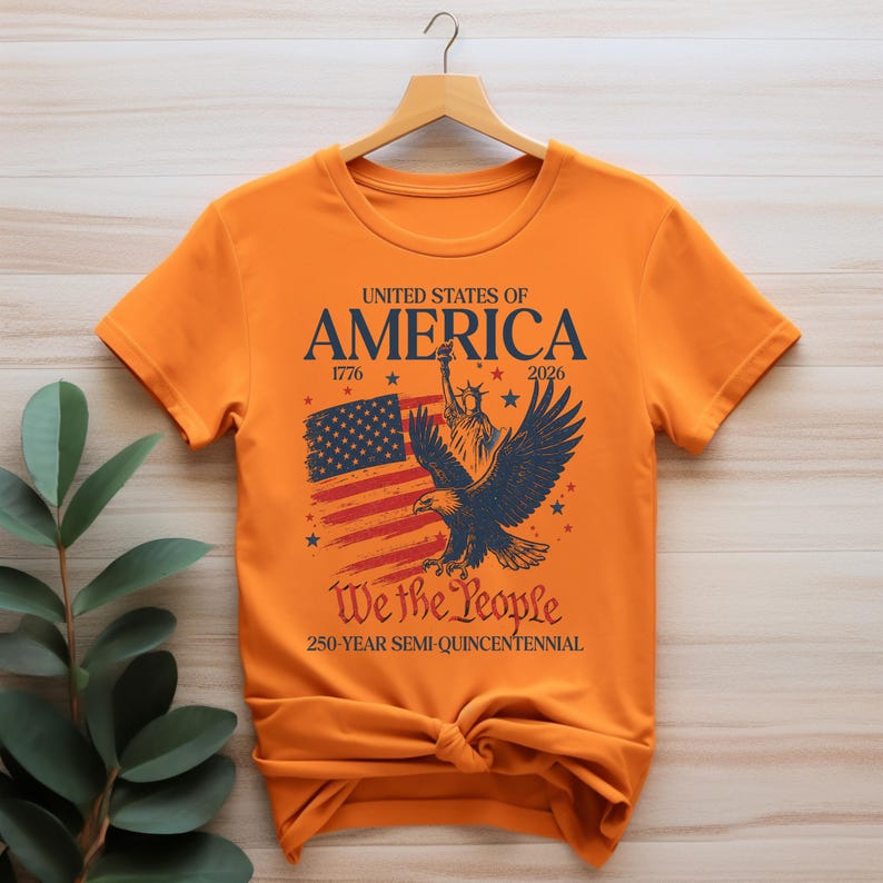 May include: Orange t-shirt with a patriotic design. The graphic includes the text "United States of America" with the years 1776 and 2026, an American flag, the Statue of Liberty, and an eagle. The phrase "We the People" is also displayed.