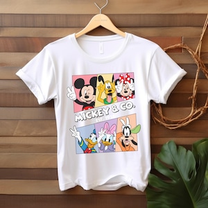 Puede incluir: Camiseta blanca con un gr&aacute;fico colorido de Mickey Mouse, Pluto, Minnie Mouse, Donald Duck, Daisy Duck y Goofy. El dise&ntilde;o incluye el texto "Mickey & Co. Est. 1928". La camiseta est&aacute; colgada en una percha de madera.