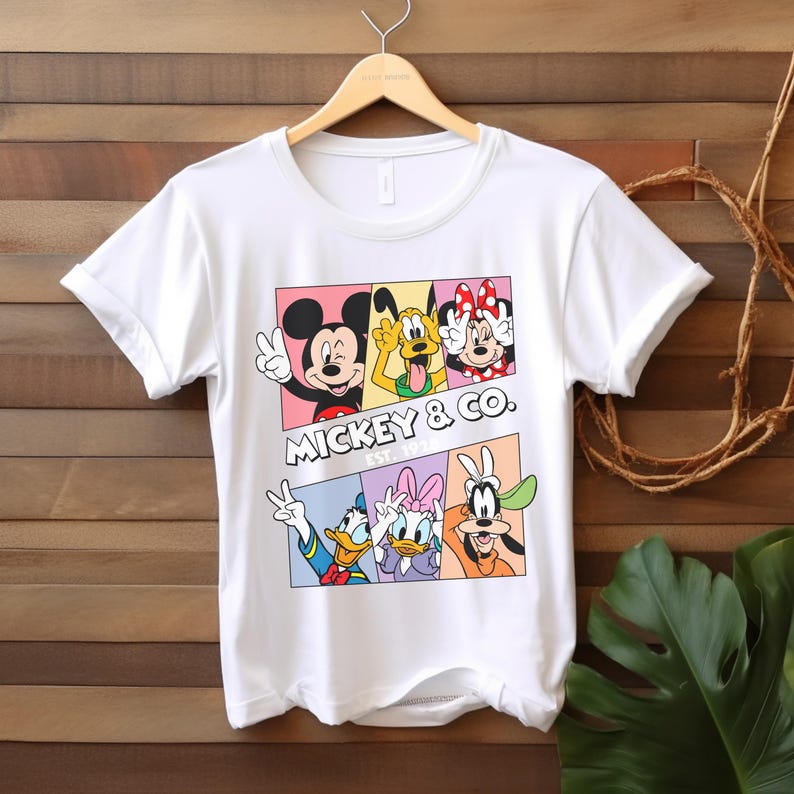 Puede incluir: Camiseta blanca con un gr&aacute;fico colorido de Mickey Mouse, Pluto, Minnie Mouse, Donald Duck, Daisy Duck y Goofy. El dise&ntilde;o incluye el texto "Mickey & Co. Est. 1928". La camiseta est&aacute; colgada en una percha de madera.