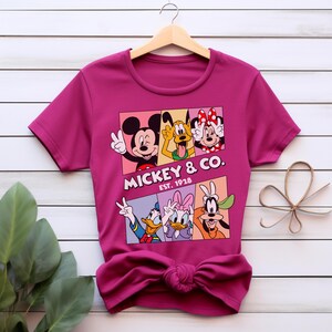 Puede incluir: Una camiseta magenta con un gr&aacute;fico de Mickey Mouse, Minnie Mouse, Pluto, Donald Duck, Daisy Duck y Goofy. El gr&aacute;fico est&aacute; en un dise&ntilde;o rectangular con el texto "Mickey & Co. Est. 1928". La camiseta est&aacute; colgada en una percha de madera.