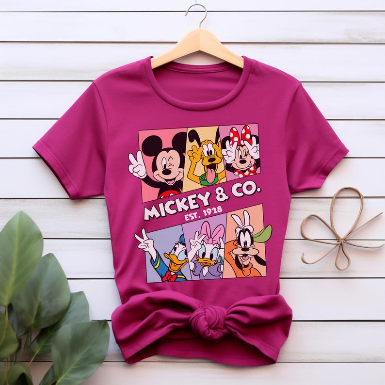 Puede incluir: Una camiseta magenta con un gr&aacute;fico de Mickey Mouse, Minnie Mouse, Pluto, Donald Duck, Daisy Duck y Goofy. El gr&aacute;fico est&aacute; en un dise&ntilde;o rectangular con el texto "Mickey & Co. Est. 1928". La camiseta est&aacute; colgada en una percha de madera.