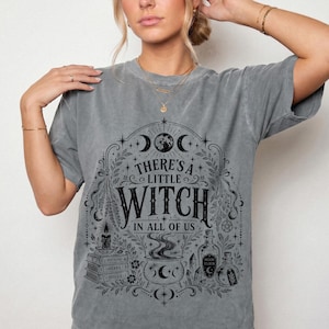 May include: A grey short-sleeve t-shirt featuring a black graphic print with the text "THERE'S A LITTLE WITCH IN ALL OF US". The design includes a moon, a stack of books labeled "POTIONS", "HERBS", and "SPELLS", a steaming cauldron, and glass bottles.