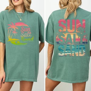 K&ouml;nnte beinhalten: Ein gr&uuml;nes T-Shirt mit den Worten "Sun Salt Sand" in einem farbenfrohen Farbverlauf. Auf der Vorderseite des T-Shirts befinden sich Palmen und ein Sonnenuntergangsdesign. Auf der R&uuml;ckseite des Shirts stehen ebenfalls die Worte "Sun Salt Sand" mit Sonnenuntergang und Palmen.