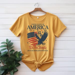 May include: Mustard yellow t-shirt with a patriotic design. The graphic includes the text "United States of America" with the years 1776 and 2026, an American flag, a bald eagle, and the words "We the People".
