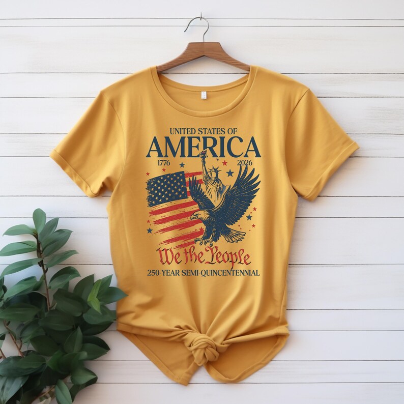 May include: Mustard yellow t-shirt with a patriotic design. The graphic includes the text "United States of America" with the years 1776 and 2026, an American flag, a bald eagle, and the words "We the People".