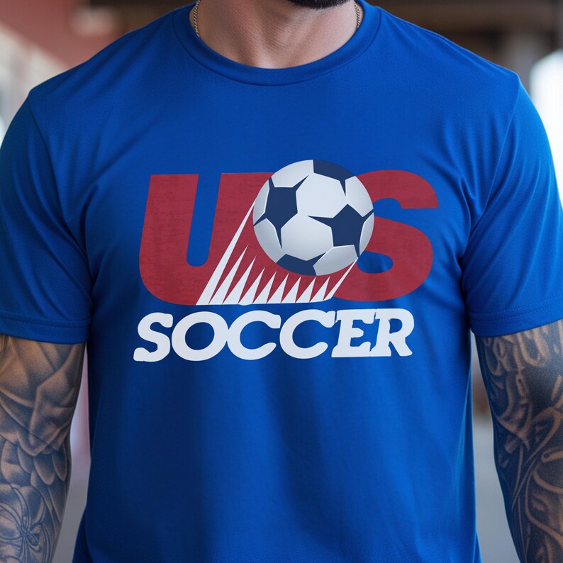 May include: A royal blue short-sleeved t-shirt featuring a graphic design with the red text US, a white and black soccer ball positioned over the letter S, and the word SOCCER printed in white capital letters underneath the graphic.