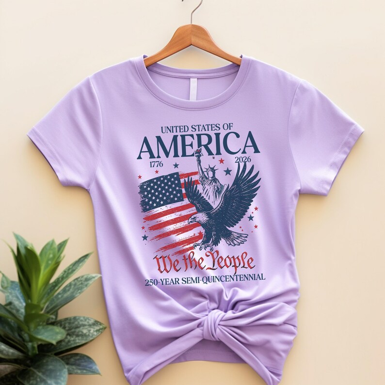 May include: A lilac t-shirt with a patriotic design. The graphic includes the text "UNITED STATES OF AMERICA" with the years 1776 and 2026, an American flag, an eagle, and the words "We the People".