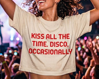May include: A cream-colored short-sleeved crop top featuring the red text KISS ALL THE TIME. DISCO, OCCASIONALLY printed on the front. The garment has a crew neckline and a straight hemline that sits above the waist, showcasing a simple design.