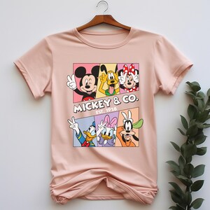 Puede incluir: Una camiseta rosa claro con un gr&aacute;fico colorido de Mickey Mouse, Pluto, Minnie Mouse, Donald Duck, Daisy Duck y Goofy. El gr&aacute;fico est&aacute; dividido en paneles e incluye el texto "Mickey & Co. Est. 1928".