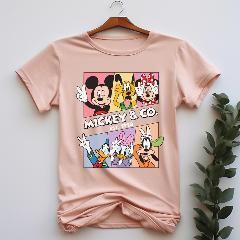 Puede incluir: Una camiseta rosa claro con un gr&aacute;fico colorido de Mickey Mouse, Pluto, Minnie Mouse, Donald Duck, Daisy Duck y Goofy. El gr&aacute;fico est&aacute; dividido en paneles e incluye el texto "Mickey & Co. Est. 1928".