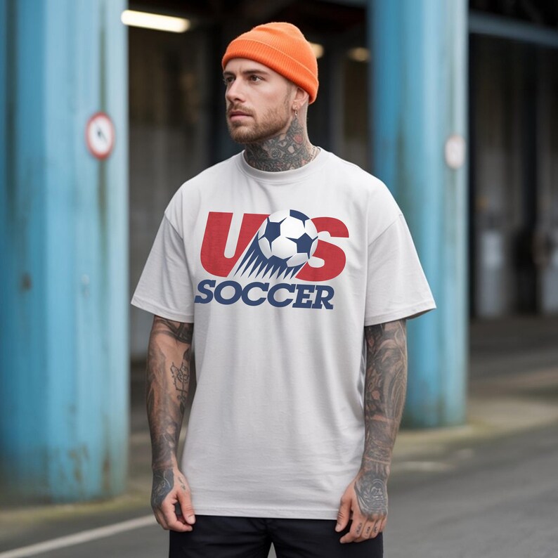 May include: A light gray short-sleeved t-shirt featuring a graphic of a soccer ball in motion over the red text US and the blue text SOCCER. The shirt is paired with an orange beanie cap, creating a casual look with a clear sports-themed design.