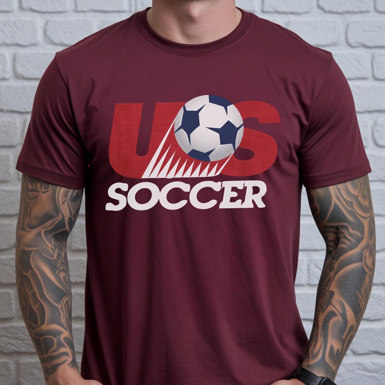 May include: A maroon short-sleeved t-shirt featuring a graphic design with the text US in large red letters, the word SOCCER in white capital letters, and a black and white soccer ball illustration with motion lines centered on the chest.