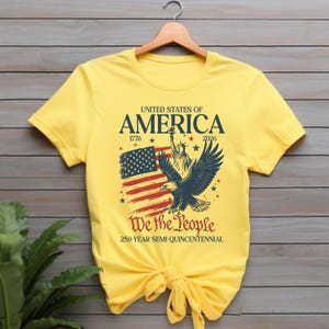 May include: A yellow t-shirt with a patriotic design. The shirt features the text "United States of America", the American flag, the Statue of Liberty, and an eagle. The text "We the People" and "250-YEAR SEMI-QUINCENTENNIAL" are also included.