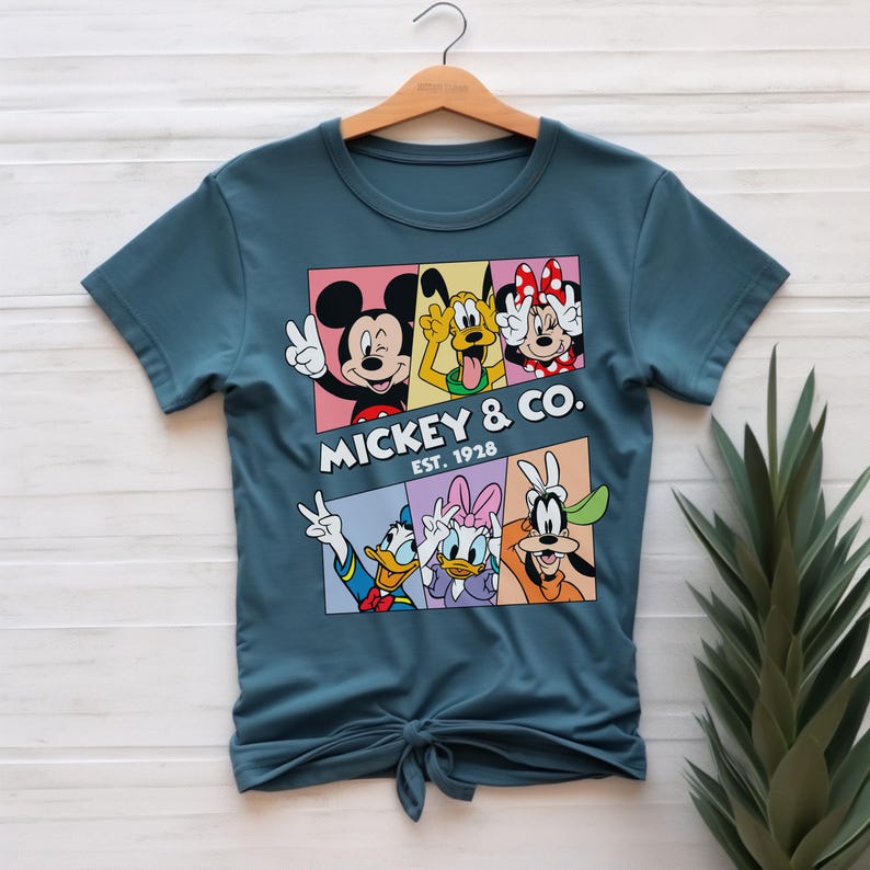 Puede incluir: Camiseta de color verde azulado con un gr&aacute;fico de Mickey Mouse, Minnie Mouse, Pluto, Donald Duck, Daisy Duck y Goofy. El gr&aacute;fico est&aacute; dividido en paneles coloridos con el texto "Mickey & Co. Est. 1928". La camiseta est&aacute; anudada en la parte inferior.
