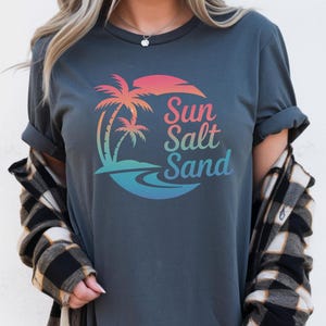 K&ouml;nnte beinhalten: Dunkelgraues T-Shirt mit einer farbenfrohen Grafik von Palmen, einer Sonne und den Worten "Sun Salt Sand". Das Design weist einen Farbverlauf aus Pink, Orange, Blau und Gr&uuml;n auf. Das Shirt hat hochgekrempelte &Auml;rmel.