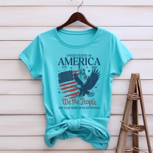 May include: A turquoise t-shirt with a patriotic design. The graphic includes the text "UNITED STATES OF AMERICA" with the years 1776 and 2026, an American flag, an eagle, and the words "We the People".