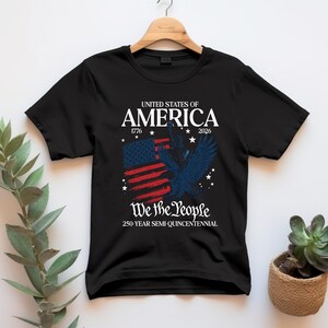 May include: Black t-shirt with a patriotic design. The design features the text "UNITED STATES OF AMERICA 1776-2026" above a distressed American flag and a blue eagle. Below the flag is the text "We the People" and "250-YEAR SEMI-QUINCENTENNIAL."