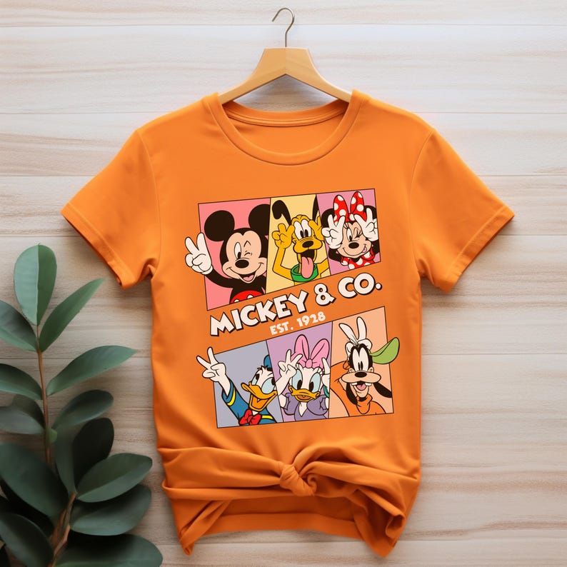 Puede incluir: Camiseta naranja con un dise&ntilde;o de Mickey Mouse y sus amigos. El dise&ntilde;o incluye a Mickey, Pluto, Minnie, Donald, Daisy y Goofy, con el texto "Mickey & Co. Est. 1928". Los personajes est&aacute;n en cuadrados coloridos.