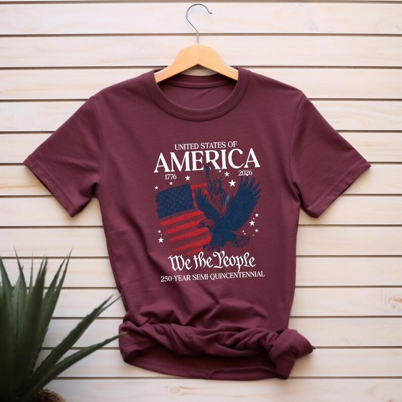 May include: A maroon t-shirt with a patriotic design. The graphic includes the text "UNITED STATES OF AMERICA 1776 2026" above an illustration of an American flag and a blue eagle. Below the image is the text "We the People 250-YEAR SEMI-QUINCENTENNIAL."