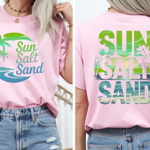 K&ouml;nnte beinhalten: Hellrosa T-Shirt mit den Worten "Sun Salt Sand" in Gr&uuml;n und Blau, mit einer Palmen-Grafik. Auf der R&uuml;ckseite des Shirts stehen ebenfalls die Worte "Sun Salt Sand". Das Shirt wird mit blauen Jeansshorts kombiniert.