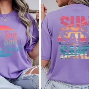 K&ouml;nnte beinhalten: Ein lilafarbenes T-Shirt mit dem Schriftzug "Sun Salt Sand" in einem Farbverlauf und einem Palmenmotiv auf der Vorderseite. Auf der R&uuml;ckseite des Shirts befindet sich derselbe Text und ein Sonnenuntergangsdesign.