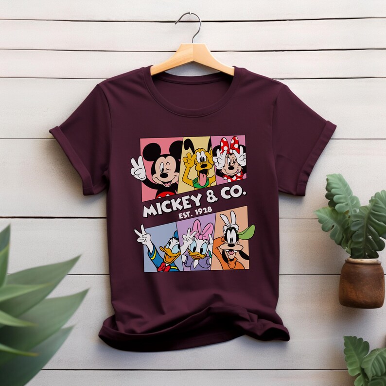 Puede incluir: Camiseta burdeos con un gr&aacute;fico colorido de Mickey Mouse, Pluto, Minnie Mouse, Donald Duck, Daisy Duck y Goofy. El gr&aacute;fico incluye el texto "Mickey & Co. Est. 1928". La camiseta est&aacute; colgada en una percha de madera.